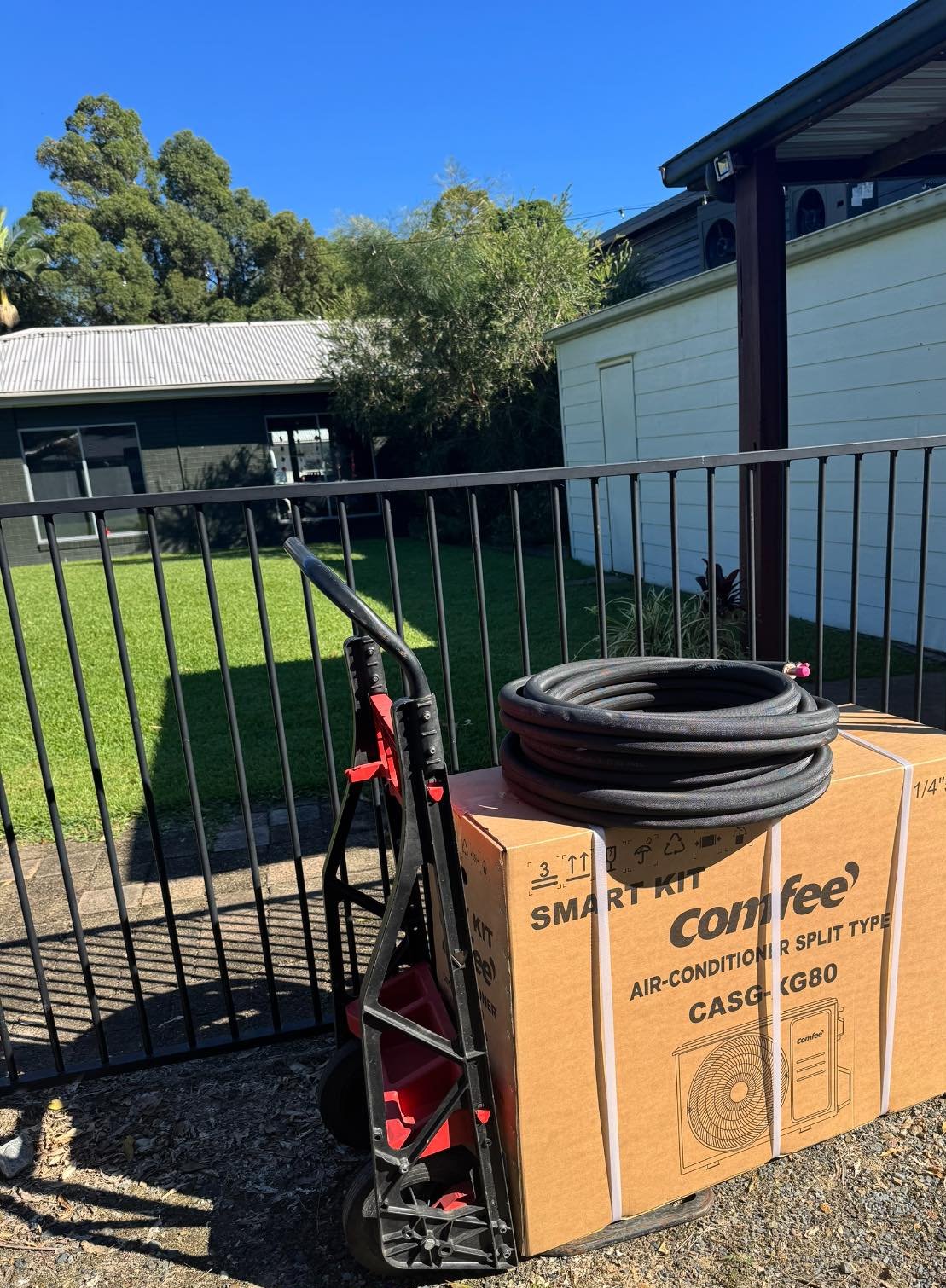 A new split system air conditioner in its box on a hand trolley, with a coil of pipe, in a backyard ready to install