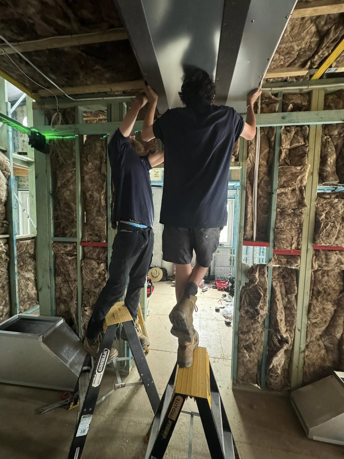 Two technicians on step ladders fitting a ducted plenum into a framed ceiling during a renovation