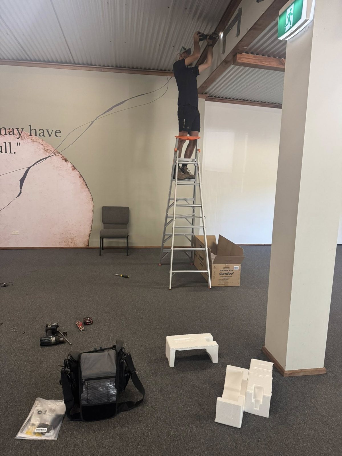 Technician up a ladder fitting an indoor unit in a large commercial room, with the new unit's box on the floor
