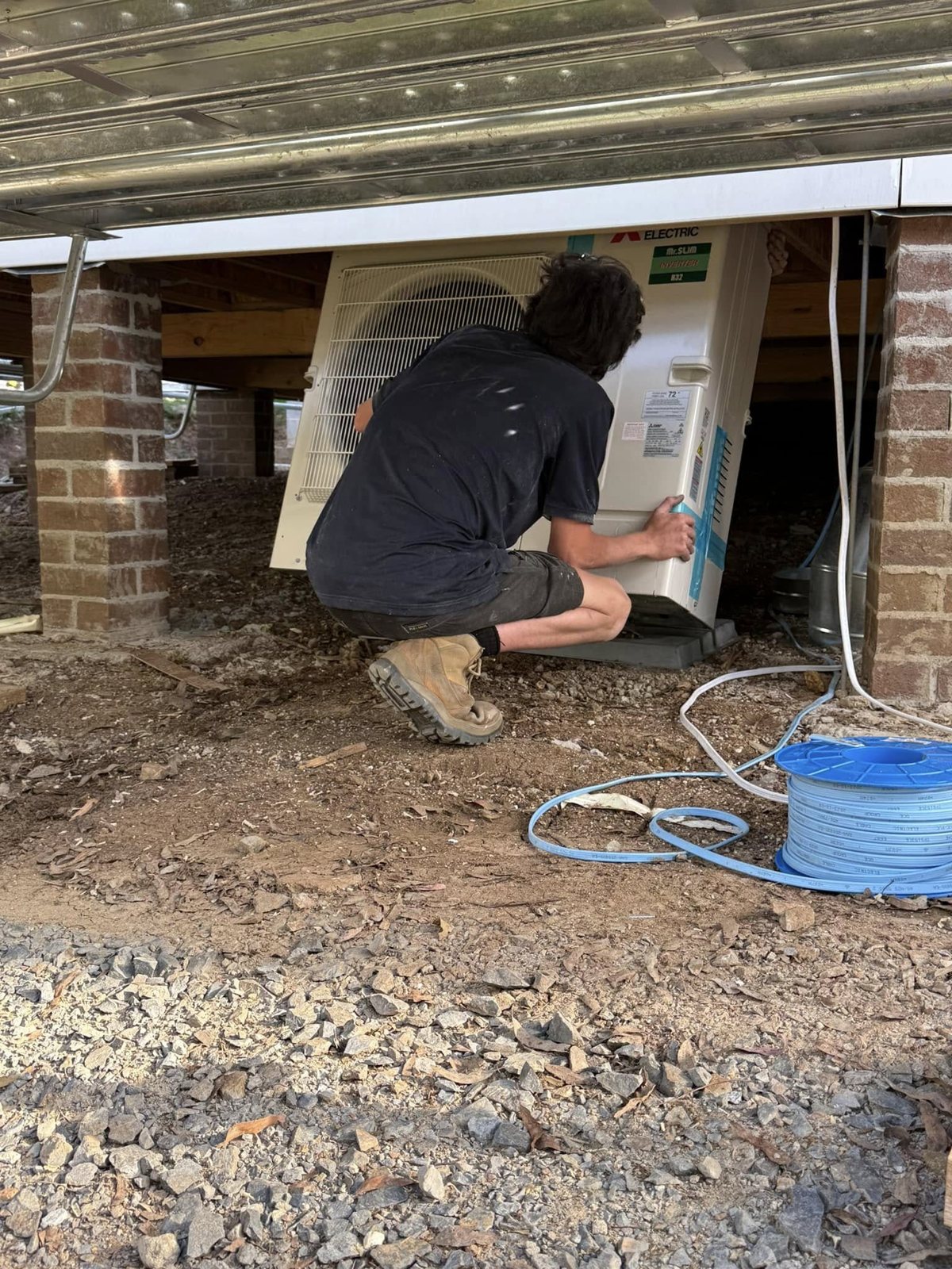 Technician crouched under a house, fitting a Mitsubishi Electric outdoor unit in a tight subfloor