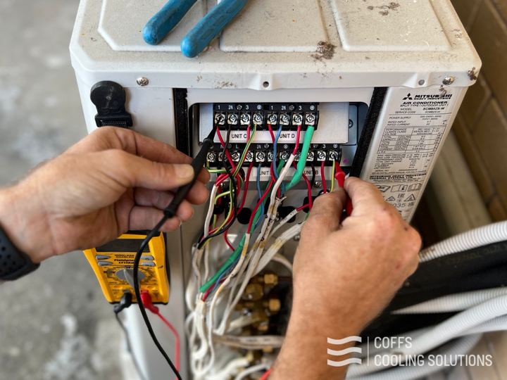 Hands testing the terminal block of a Mitsubishi Heavy Industries outdoor unit with a multimeter